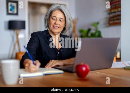 Smiling mature businesswoman writing notes in notebook while working remotely using laptop computer at home office Stock Photo