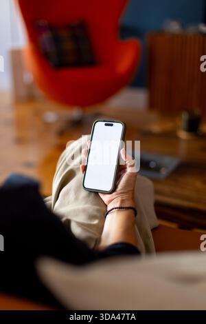 Woman sitting on comfortable sofa at home, holding smartphone with blank white screen, perfect for mockup or app design presentation Stock Photo