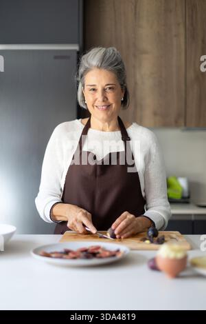 Senior woman wearing an apron and smiling, preparing fresh figs on a cutting board in a modern kitchen Stock Photo