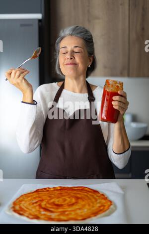 Woman in kitchen smelling delicious tomato sauce while preparing homemade pizza, representing passion for cooking and food Stock Photo