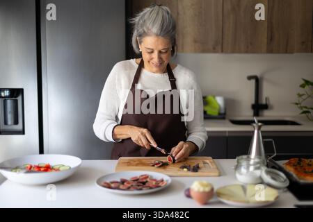 Happy mature woman wearing an apron preparing healthy food, cutting figs on a cutting board, enjoying cooking at home Stock Photo