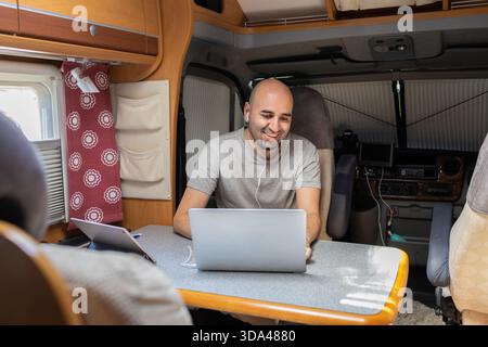 A man using a laptop and wearing earphones, engaged in remote work inside a cozy motorhome, demonstrating the flexibility of telecommuting while trave Stock Photo