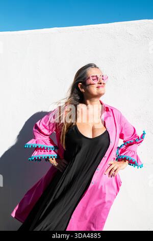 Stylish young woman posing in sunlight wearing pink heart-shaped sunglasses, black dress, and pink ruffled overshirt, standing against a white wall. Stock Photo