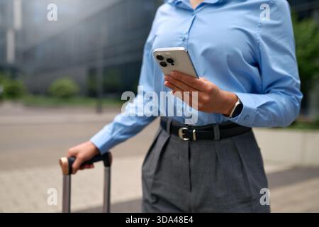 Close-up shot of a woman in blue shirt using smartphone outdoors, holding suitcase handle. Business travel, modern technology, communication, professi Stock Photo