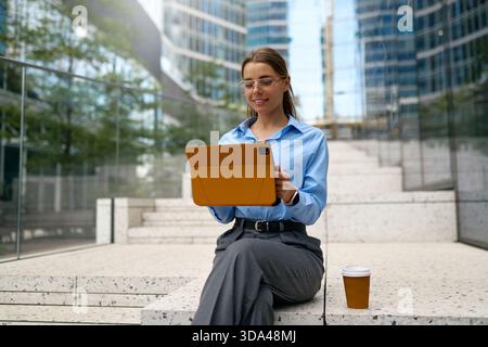 Young businesswoman sitting on outdoor steps, using a tablet in an orange case, wearing glasses and a blue shirt, with a takeaway coffee cup beside he Stock Photo