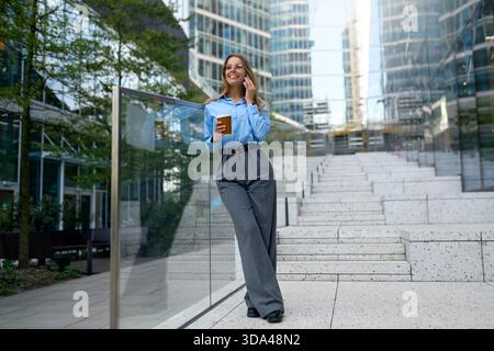 Confident young woman in blue shirt and glasses leaning on glass railing, holding coffee and talking on smartphone outdoors in modern urban business d Stock Photo