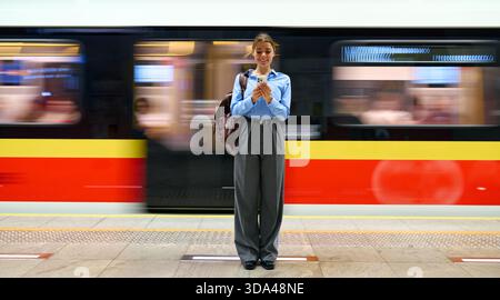 Front view of smiling young woman using smartphone while standing at metro station with fast-moving train in background. Concept of urban commute, tec Stock Photo