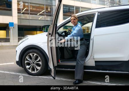 Smiling businesswoman in blue shirt getting into a white car in a modern city. Concept of travel, commuting, transportation, professional lifestyle, a Stock Photo