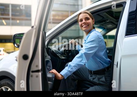 Smiling businesswoman in blue shirt getting into a white car in a modern city. Concept of travel, commuting, transportation, professional lifestyle, a Stock Photo
