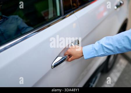 Close-up of female hand in blue shirt reaching for white car door handle. Modern vehicle, city background. Reflected smiling woman seen in car window. Stock Photo