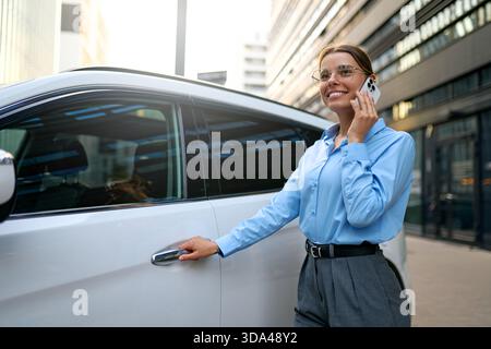 Happy businesswoman in blue shirt chatting on phone, leaning on white car in urban setting. Professional woman enjoying conversation, modern city life Stock Photo