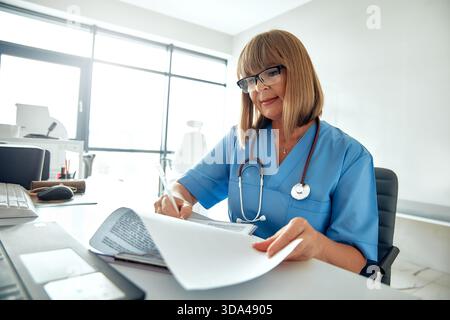 A focused female doctor in scrubs meticulously reviews medical documents at her modern office desk, embodying professionalism and care Stock Photo