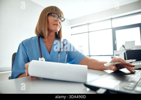 A highly focused and dedicated female doctor dressed in blue scrubs diligently reviews important medical documents while working on her laptop in a mo Stock Photo