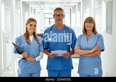 A dedicated healthcare team of three smiling individuals in bright blue scrubs is ready to assist patients in a welcoming hospital hallway, radiating Stock Photo