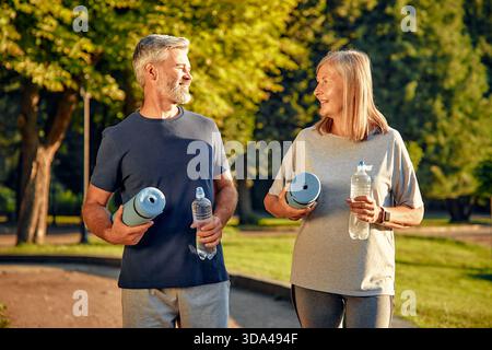 A fit and happy mature couple is enjoying a beautiful sunny day outdoors in a lovely park, happily holding their yoga mats and water bottles while eng Stock Photo