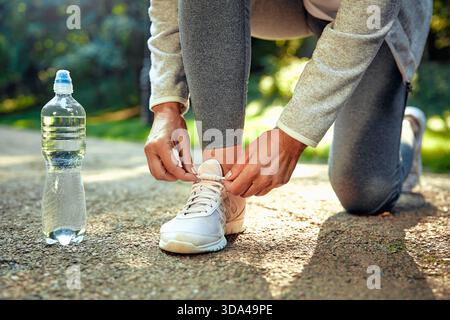 A person is kneeling outside in nature, diligently tying their running shoe, with a water bottle conveniently placed nearby, perfectly illustrating a Stock Photo