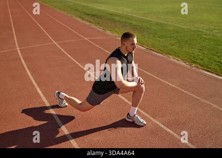 Athletic man in black tank top performing lunge stretch on red running track under sunlight. Concept: flexibility, warmup, outdoor fitness, strength, Stock Photo