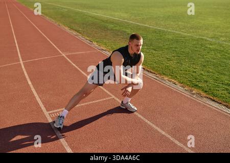 Sporty young man in black sleeveless shirt stretching legs before running on stadium track. Concept: warmup, flexibility, sport, strength, preparation Stock Photo