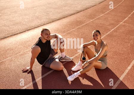 Young athletic man and woman sitting on the track, smiling and chatting after training. He holds a smartphone, she holds a water bottle. Concept: spor Stock Photo