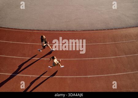 Drone shot of a man and woman running on a curved red track, casting long shadows in evening light. Concept of fitness, teamwork, and determination. Stock Photo