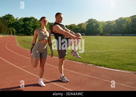 Young athletic man and woman in sportswear stretching on running track under the morning sun. Concept of fitness, warm-up, and healthy lifestyle. Stock Photo