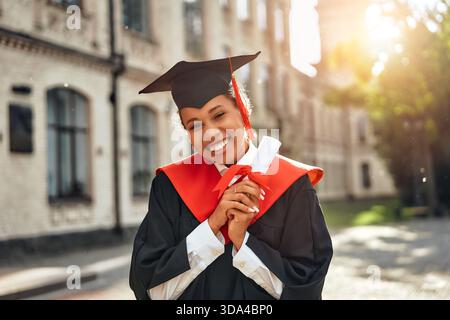 Under the bright sunshine, a young woman proudly shows her diploma in front of a magnificent university building, celebrating her academic success on Stock Photo