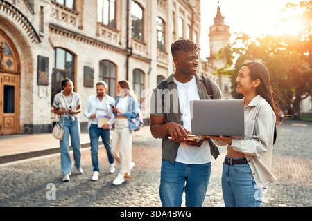 Two college students are outside collaborating on a laptop while two others walk and chat near campus buildings in the background, showcasing a scene Stock Photo