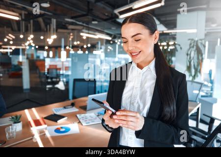 Business woman in professional attire using a smartphone in a modern office environment, emphasizing the integration of technology in workplace. Stock Photo