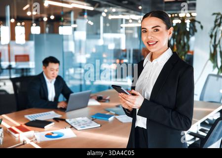 Two professional business workers in formal suits using phone and laptop, sitting in office and using modern technology. Stock Photo