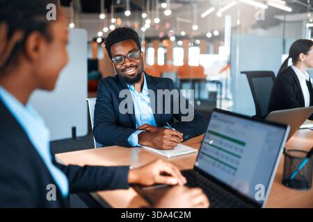 A team of young business workers in formal suits working with laptops while sitting in a modern office. Employees discussing a work plan or project on Stock Photo