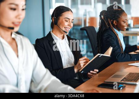 Team of successful call center employees with headsets sitting with laptops at the table and answering calls, providing assistance and support to cust Stock Photo