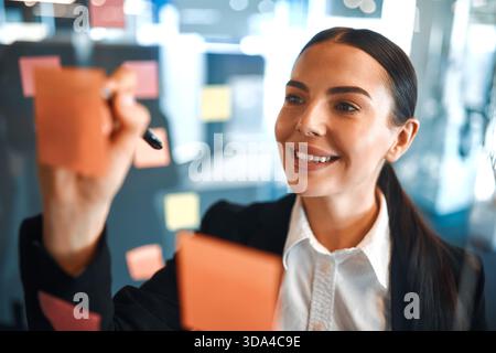Young business woman in a formal suit posting sticky notes with sticky notes sticking them on glass, reminders of creative brainstorming in a modern c Stock Photo