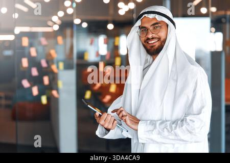 Young Middle Eastern corporate businessman in traditional Emirati clothing using a tablet in the office. Business team working and brainstorming in UA Stock Photo