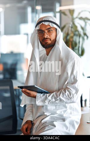 Young Middle Eastern corporate businessman in traditional Emirati clothing using a tablet in the office. Business team working and brainstorming in UA Stock Photo