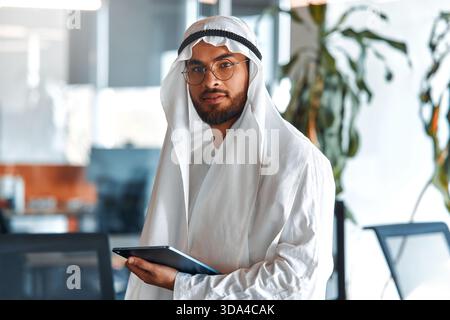 Young Middle Eastern corporate businessman in traditional Emirati clothing using a tablet in the office. Business team working and brainstorming in UA Stock Photo