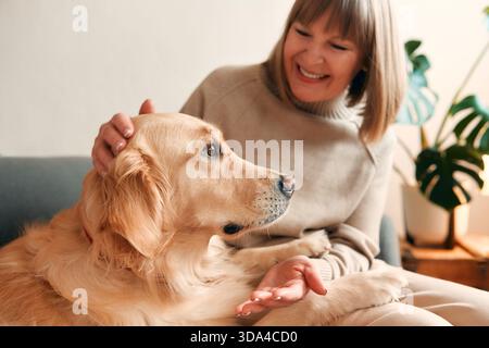 Beautiful aged woman sitting on sofa with her dog in cozy living room and having fun. Stock Photo