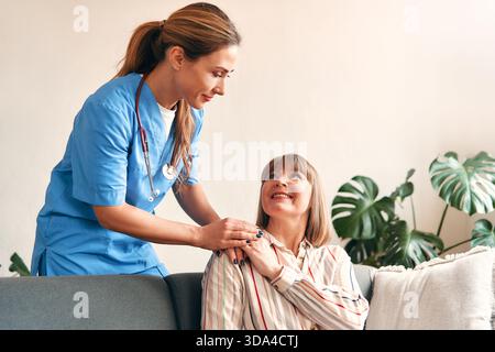 Happy young female nurse provide care medical service help support smiling mature woman at homecare medical visit, lady carer doctor give empathy enco Stock Photo