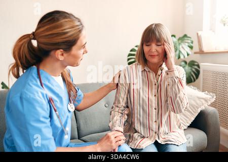 A female family doctor at home in a medical uniform with a stethoscope examining an elderly woman who complains of a headache while sitting on the sof Stock Photo