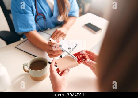 Female doctor in medical uniform with stethoscope sitting at table with laptop taking notes and talking to patient about pills in pillbox. Health care Stock Photo