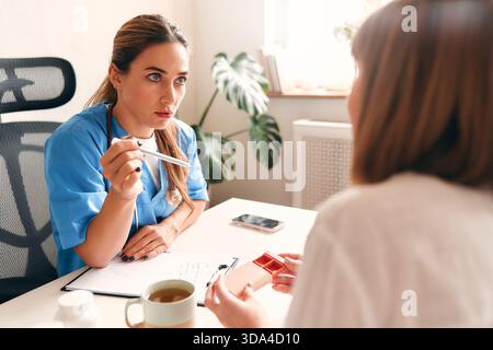 Female doctor in medical uniform with stethoscope sitting at table with laptop taking notes and talking to patient about pills in pillbox. Health care Stock Photo