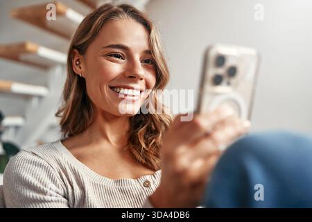 Happy relaxed young woman sitting on couch using cell phone, smiling lady laughing holding smartphone, looking at cellphone enjoying doing online ecom Stock Photo