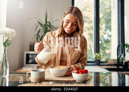 A woman preparing herself breakfast, pouring milk into a plate with cereal, strawberries and a cup of coffee standing nearby. A woman is having breakf Stock Photo