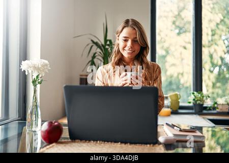 Young happy woman sitting at the table in the kitchen, working on a laptop and drinking coffee. Business woman freelancer working remotely from home. Stock Photo
