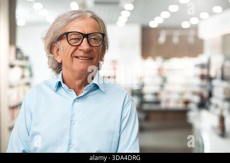 Portrait of a cheerful senior man wearing glasses and light blue shirt, smiling while standing in a modern pharmacy. Concept of health, medicine and p Stock Photo
