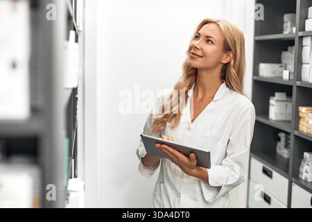 A confident female pharmacist in a white coat is using a tablet in a pharmacy storage room. Concept of modern pharmaceutical inventory management and Stock Photo