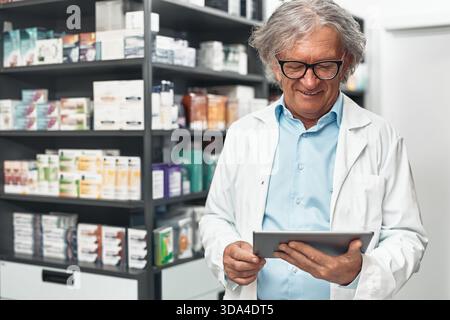 Elderly male pharmacist in white coat and glasses stands in a pharmacy, smiling while using a digital tablet against shelves with medicine and health Stock Photo