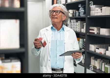 Elderly male pharmacist with gray hair and glasses in a white lab coat checking medicine boxes on pharmacy shelves while holding a digital tablet. Con Stock Photo
