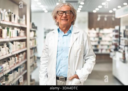 Confident senior pharmacist in a white coat and blue shirt standing in a pharmacy aisle, smiling and looking at camera. Concept of healthcare and prof Stock Photo