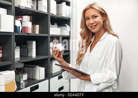 Smiling female pharmacist with tablet checks medicine inventory in pharmacy storage. Concept: pharmacy work. Stock Photo