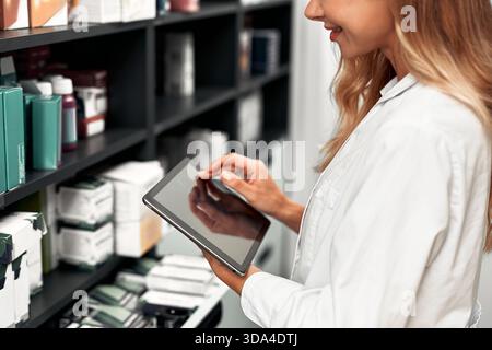 A smiling female pharmacist in a white coat uses a digital tablet while organizing medication boxes in a modern pharmacy. Concept of pharmaceutical in Stock Photo